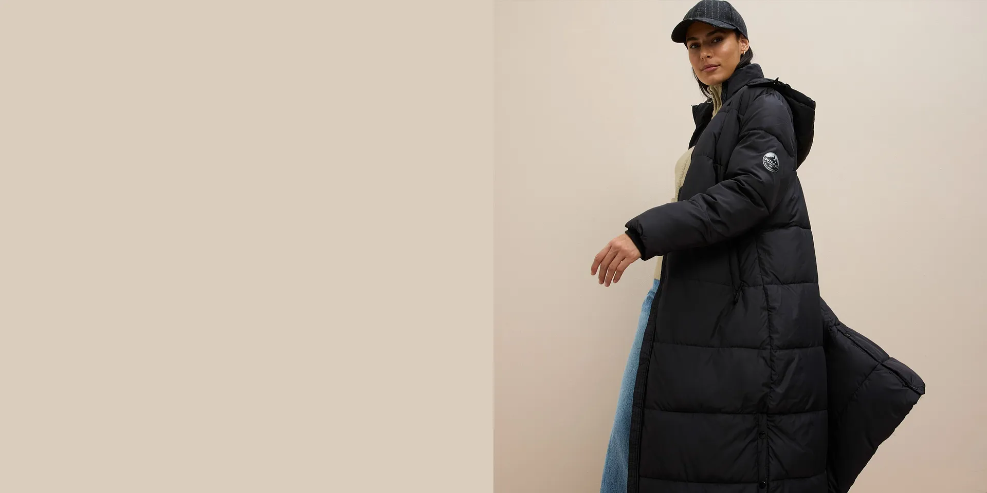 Lång svart jacka över beige tröja, vida jeans och mössa fulländar looken.