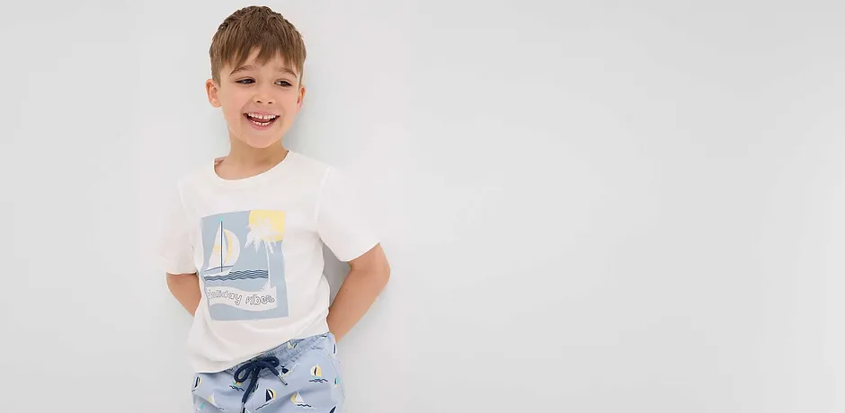 Ett glatt barn i en vit t-shirt och ljusblå shorts, båda med segelbåtsmönster.