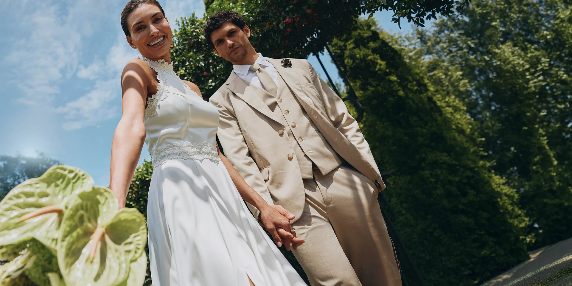 Elegant brudpar i solsken: hon i vit kl&auml;nning, han i beige kostym med solglas&ouml;gon.