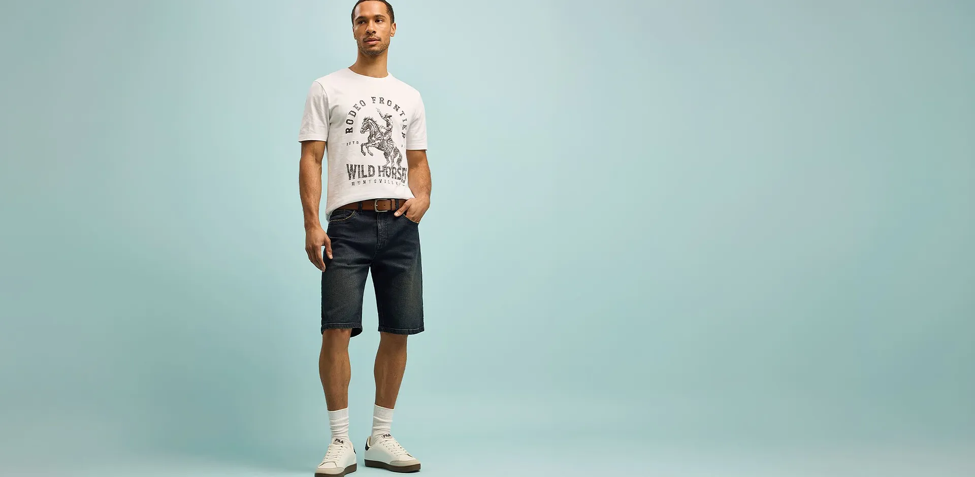 Man b&auml;r vit T-shirt med tryck och m&ouml;rka shorts i en avslappnad pose.

