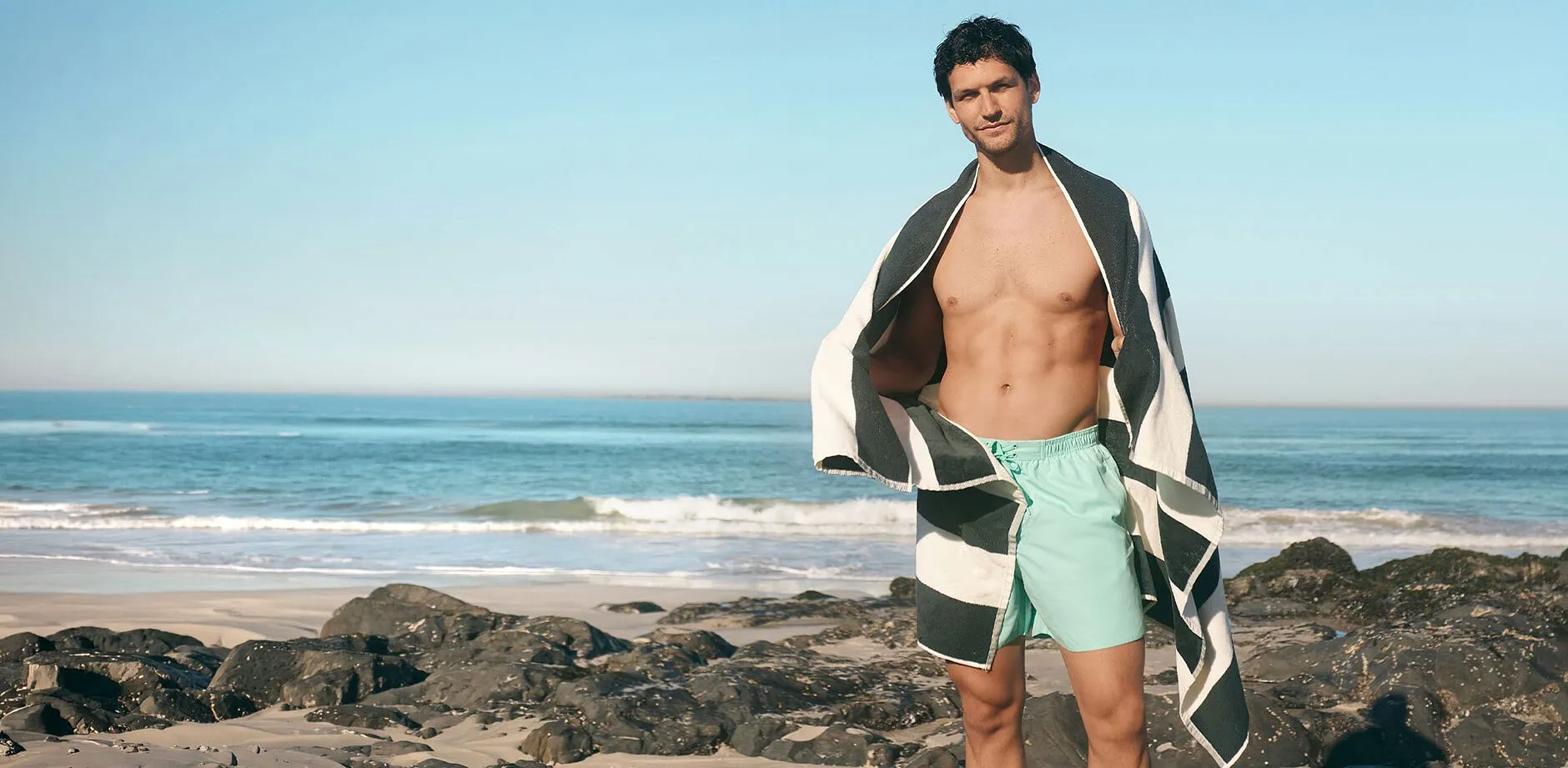 En man b&auml;r mintgr&ouml;na badshorts och en stilren randig strandhandduk &ouml;ver axlarna.