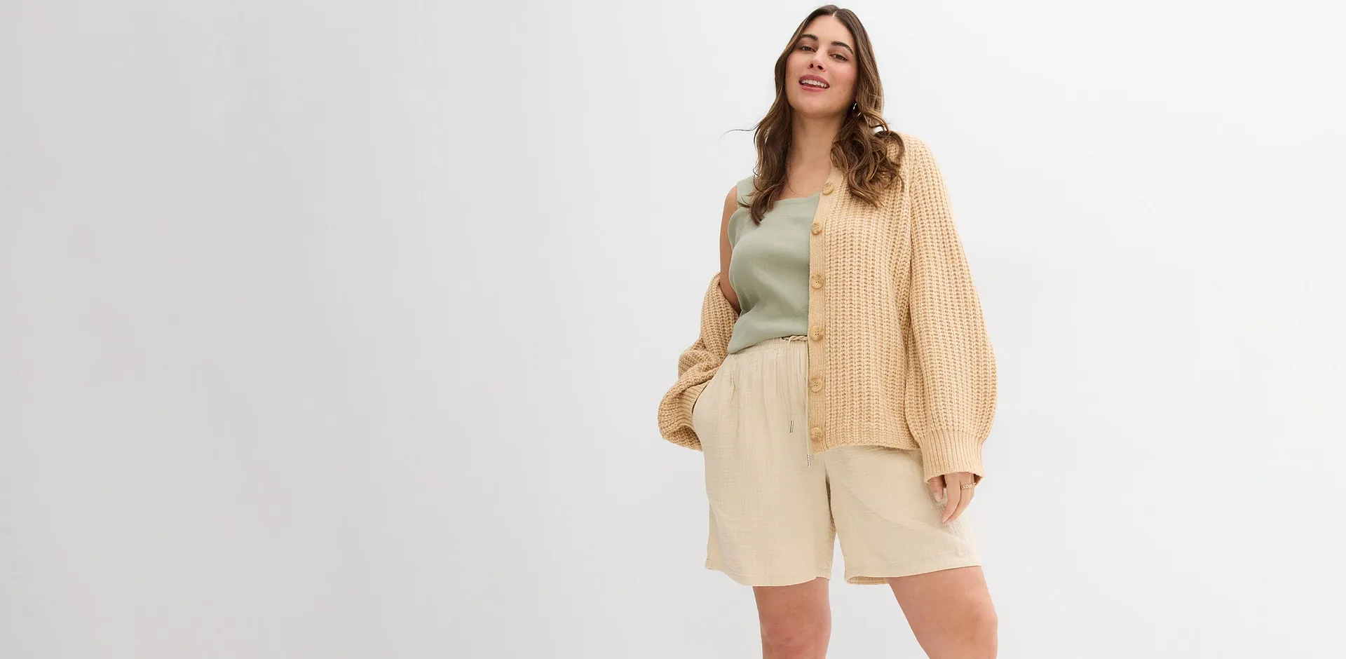 Kvinna b&auml;r beige cardigan, ljusgr&ouml;n topp och beige shorts.

