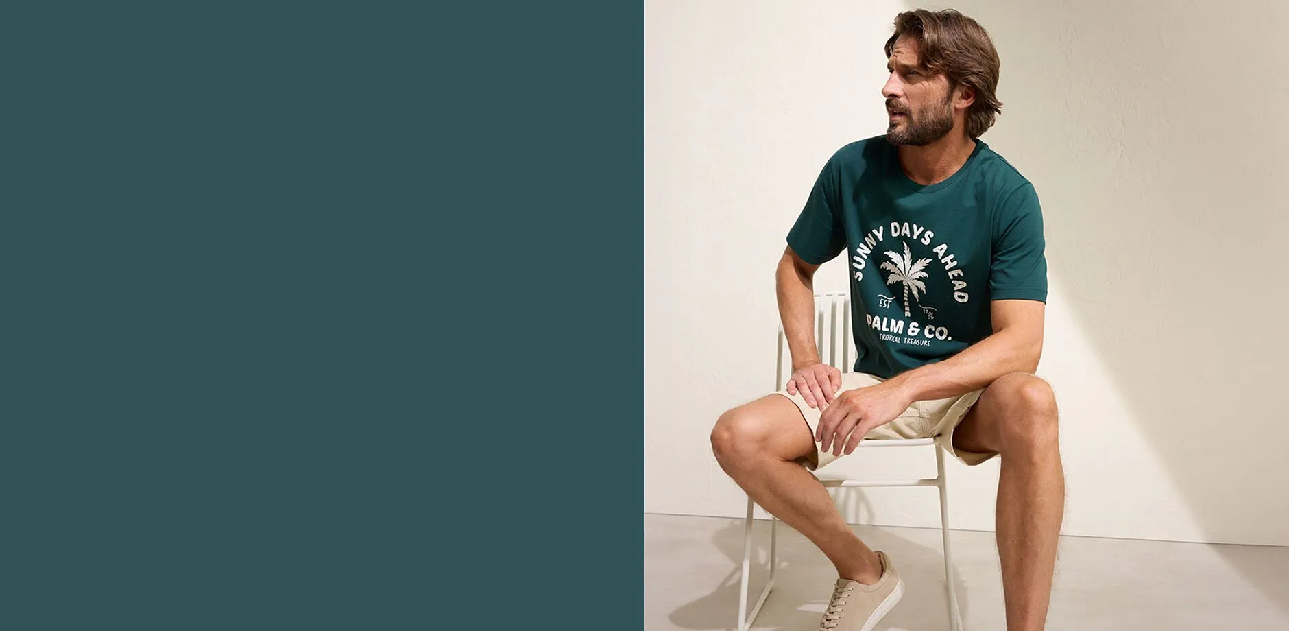 En man b&auml;r en gr&ouml;n t-shirt med palmtryck, beige shorts och vita sneakers.
