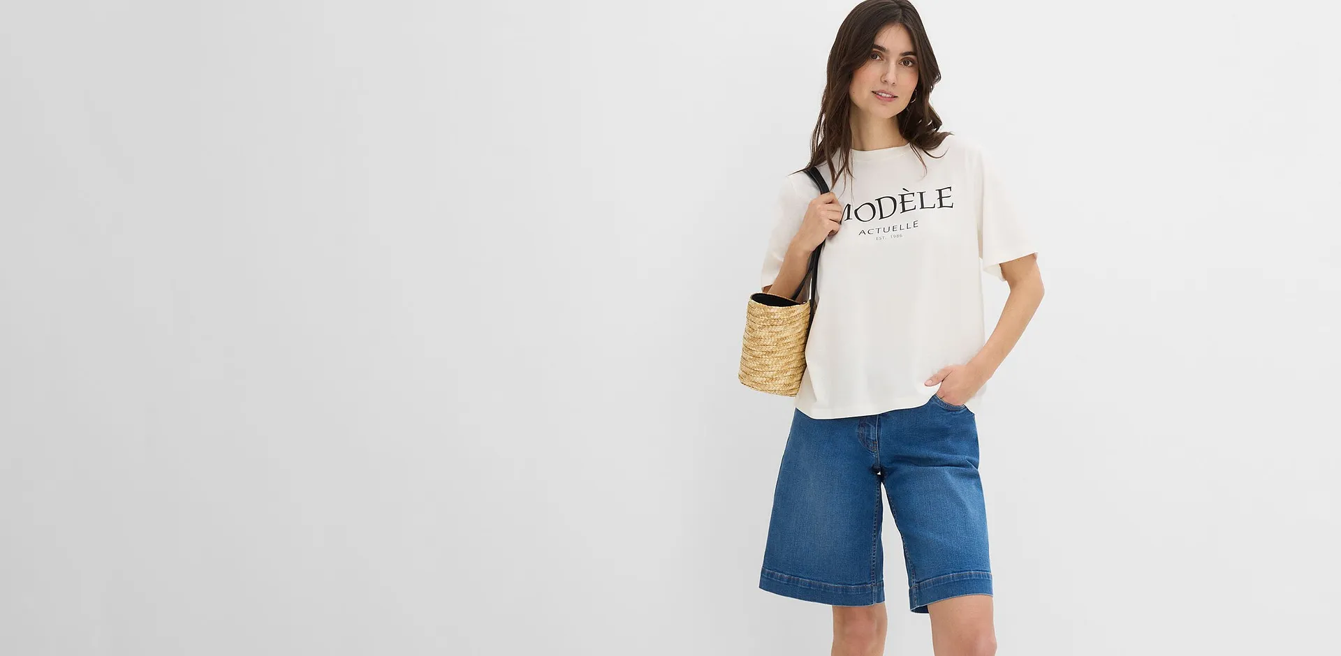 Hon b&auml;r en avslappnad vit T-shirt med texttryck och vida bl&aring; jeansbermudashorts.