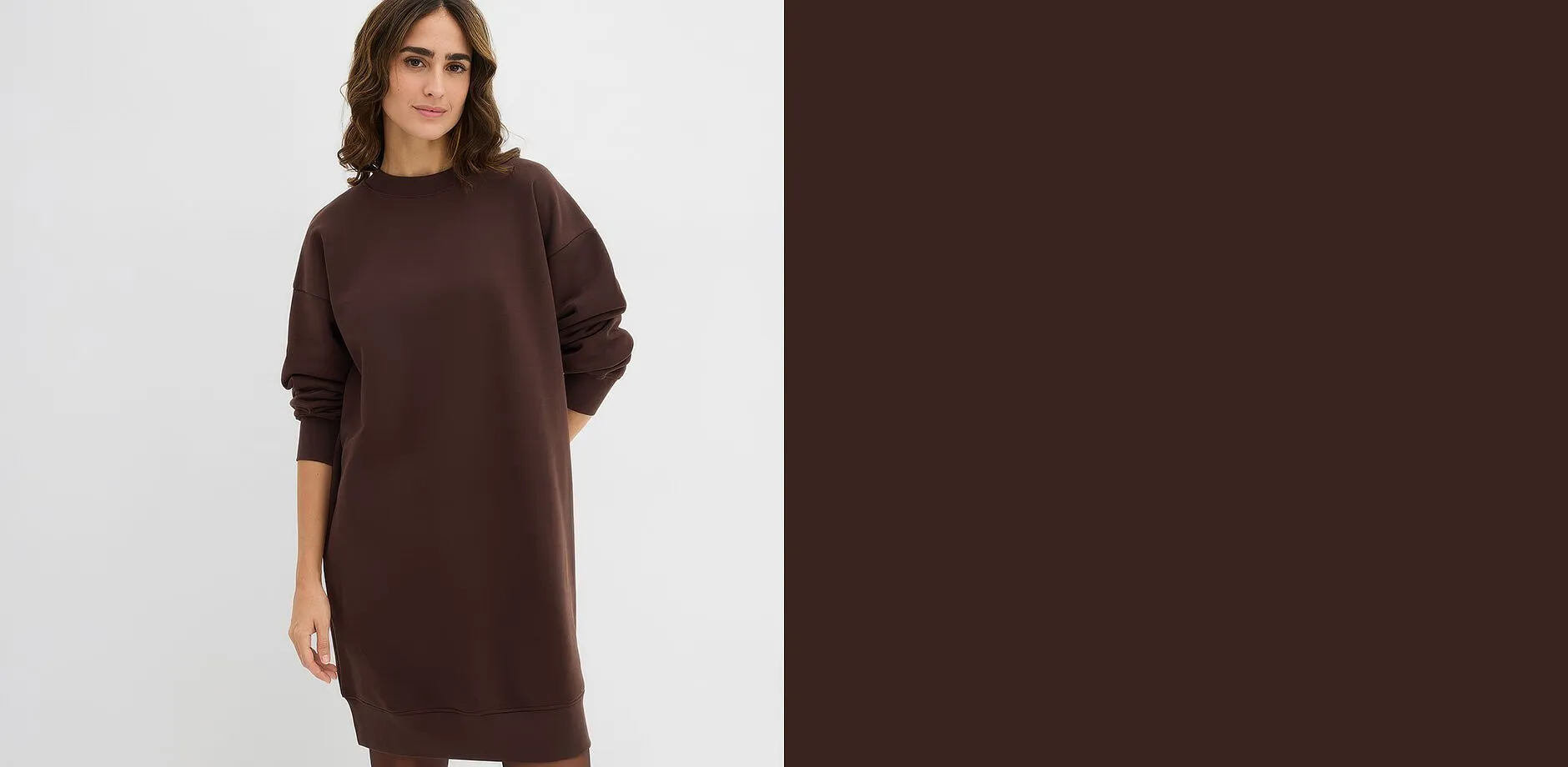 En kvinna har p&aring; sig en l&aring;ng, l&ouml;st sittande m&ouml;rkbrun sweatshirtkl&auml;nning.