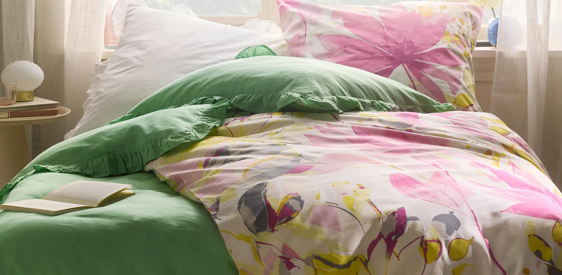 F&auml;rgglada s&auml;ngkl&auml;der med blomm&ouml;nster, kombinerat med gr&ouml;n kudde. Skira gardiner ramar in s&auml;ngen.