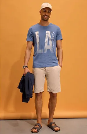 Blå T-shirt med ”LA”-tryck, ljusa shorts, vit keps och tofflor. Avslappnad sommarlook.