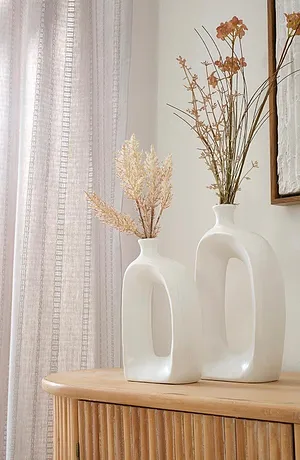 Ljusa, luftiga gardiner ramar in eleganta vita vaser med torkade blommor på en träskåp.