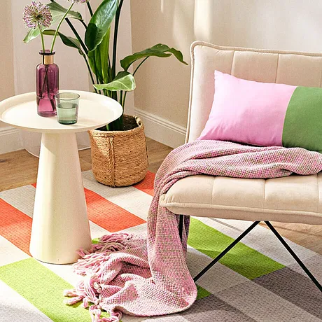 Ljusa textilier i pastellfärger: rosa filt, gröna och rosa kuddar på en beige fåtölj.