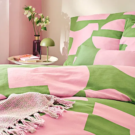 Fräscha sängkläder i rosa och grönt med mönster av geometriska former. En vävd filt fulländar looken.