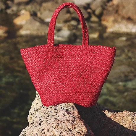 Ljusröd handväska av strå vilar på stenar vid vattnet, perfekt för sommarens strandutflykter.