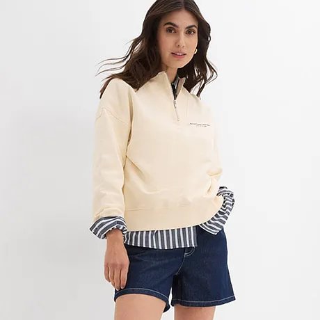 En naturfärgad tröja över en randig skjorta, kombinerad med jeansshorts.