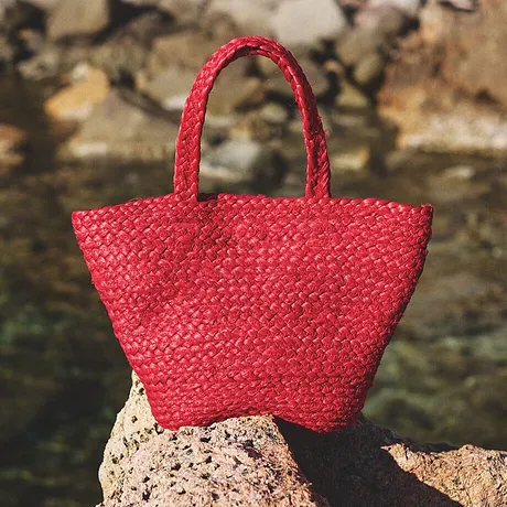 Klarröd handväska i strålook vilar på stenar vid vattnet, perfekt för sommarens strandutflykter.