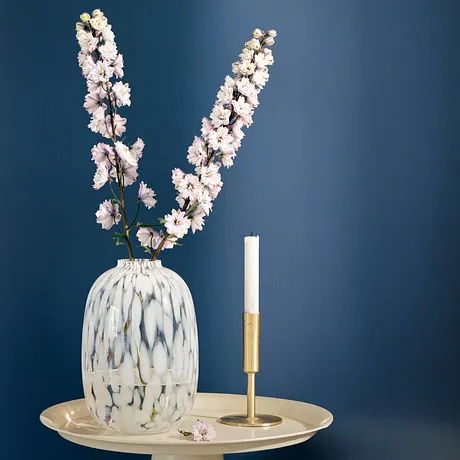 Elegant vit vas med späd körsbärsblom på en guldfärgad bricka bredvid ett smalt ljus.
