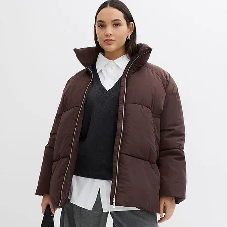 Brun vinterjacka med hög krage, under det en mörk tröja och vit skjorta.

