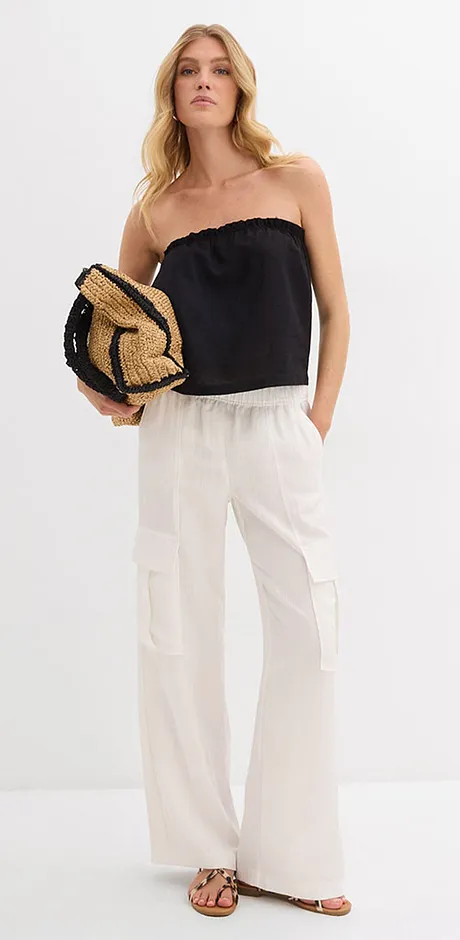 Somrig outfit med en svart off-shoulder-topp, luftiga vita byxor och en flätad väska i strålook.