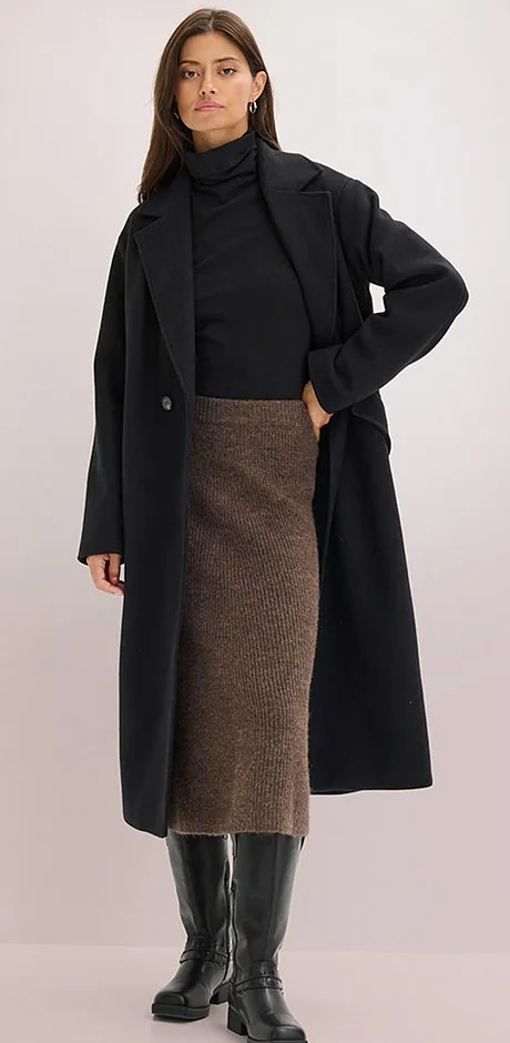 Elegant svart kappa över polotröja, kombinerad med en brun midikjol och knähöga stövlar.

