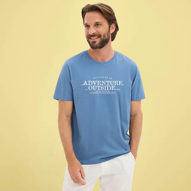 Man b&auml;r en ledig bl&aring; t-shirt med vitt tryck och ljusa byxor.

