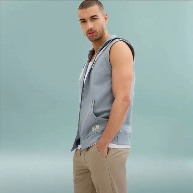 Man b&auml;r en ljusbl&aring; sweatshirtv&auml;st med huva &ouml;ver en vit skjorta och beige byxor.

