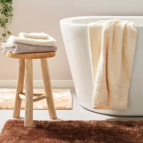 Vita handdukar i creme- och beige toner smyckar badkaret och pallen i det ljusa badrummet.