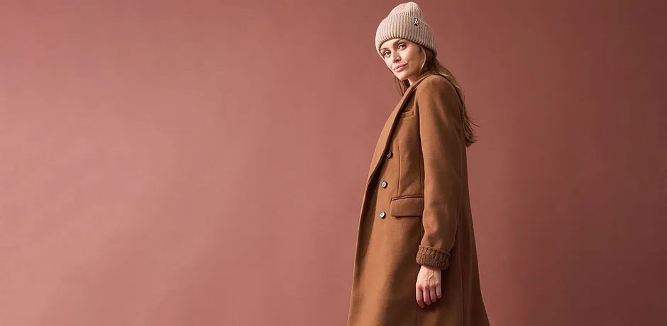 Lång kamelbrun kappa, beige stickad mössa och en enkel outfit under.

