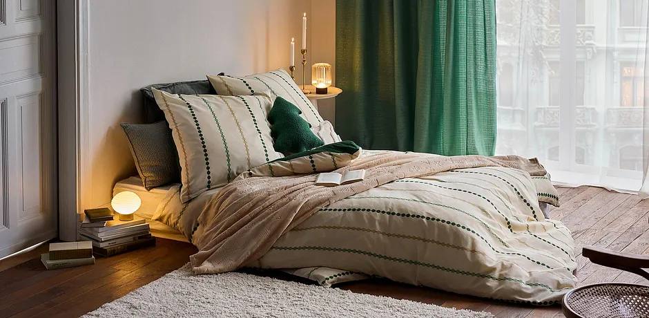 Randiga sängkläder i beige, gröna kuddar och gardiner skapar en mysig atmosfär.

