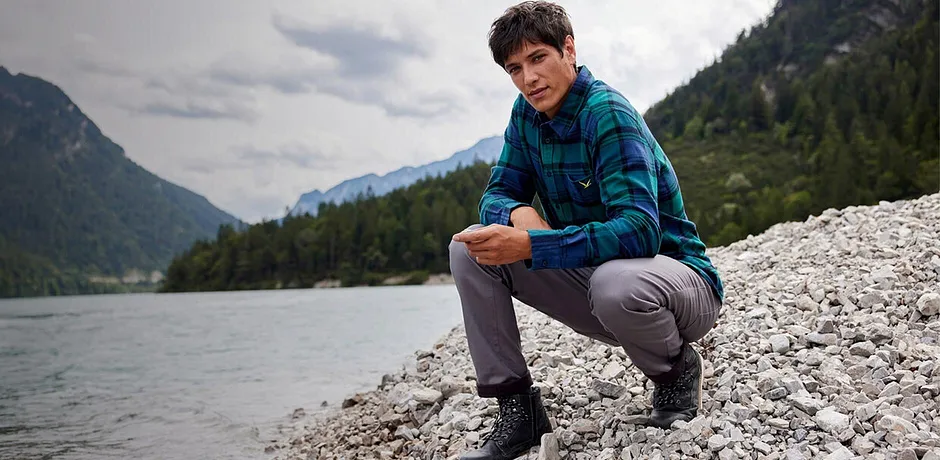 En vandrare i rutig skjorta och grå byxor sitter vid flodstranden i ett bergslandskap.

