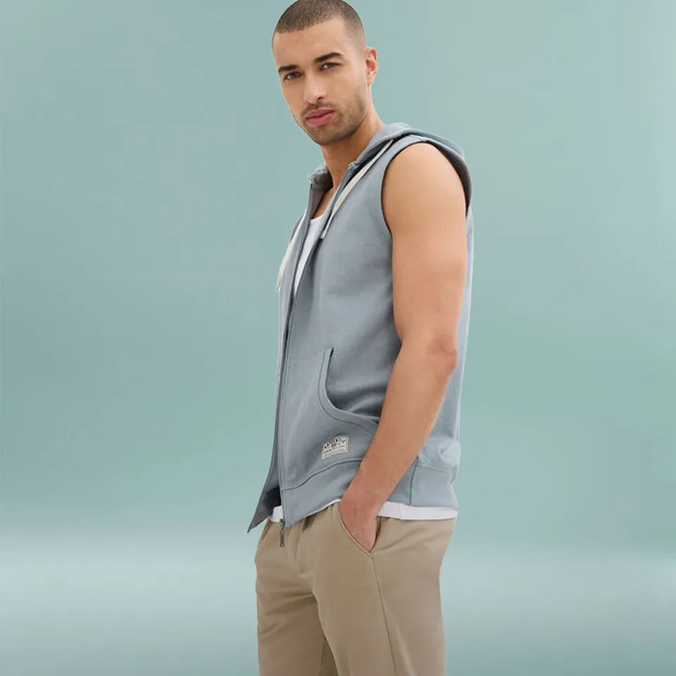 Man b&auml;r ljusbl&aring; v&auml;st i sweatmaterial med huva &ouml;ver vit t-shirt och beiga byxor.

