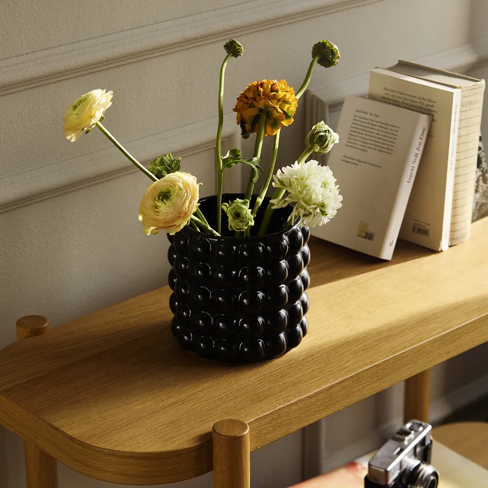 Svart vas med f&auml;rska blommor p&aring; ett tr&auml;bord bredvid b&ouml;cker, dekorativt arrangerade.

