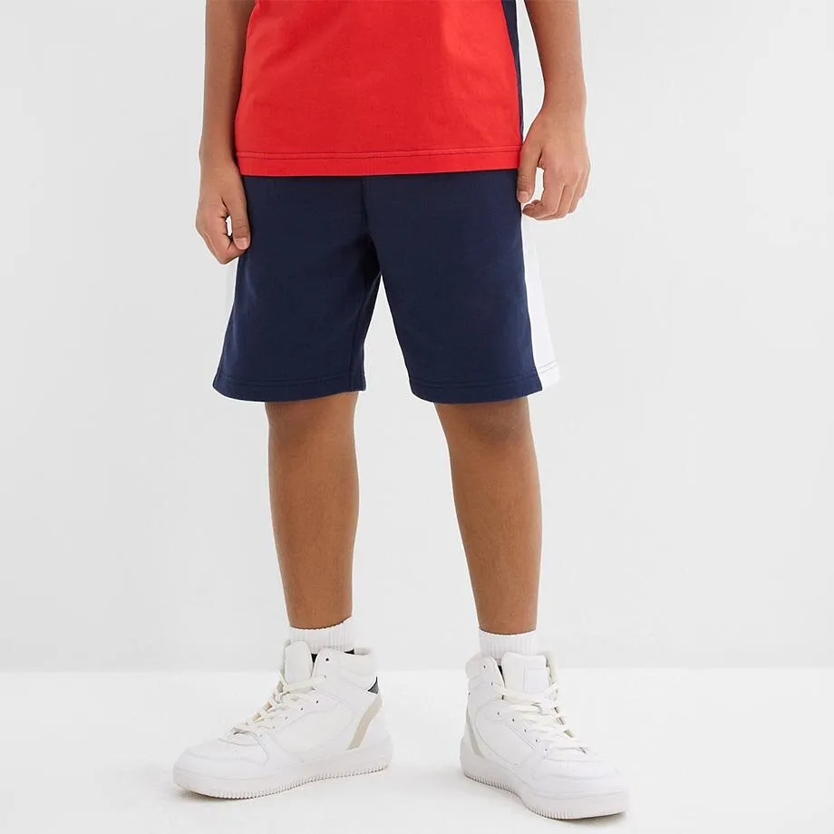 Ett barn b&auml;r en r&ouml;d &ouml;verdel och m&ouml;rkbl&aring; shorts.