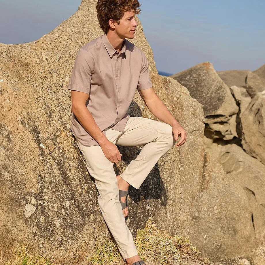 En man har p&aring; sig en beige skjorta och ljusa chinosbyxor med sandaler p&aring; stranden.

