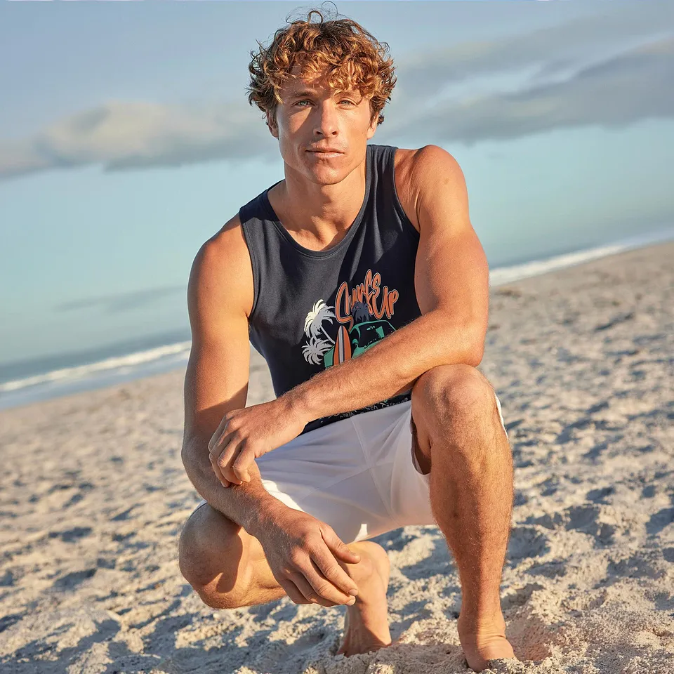 Man b&auml;r ett bl&aring;tt linne med motiv och vita shorts p&aring; stranden.

