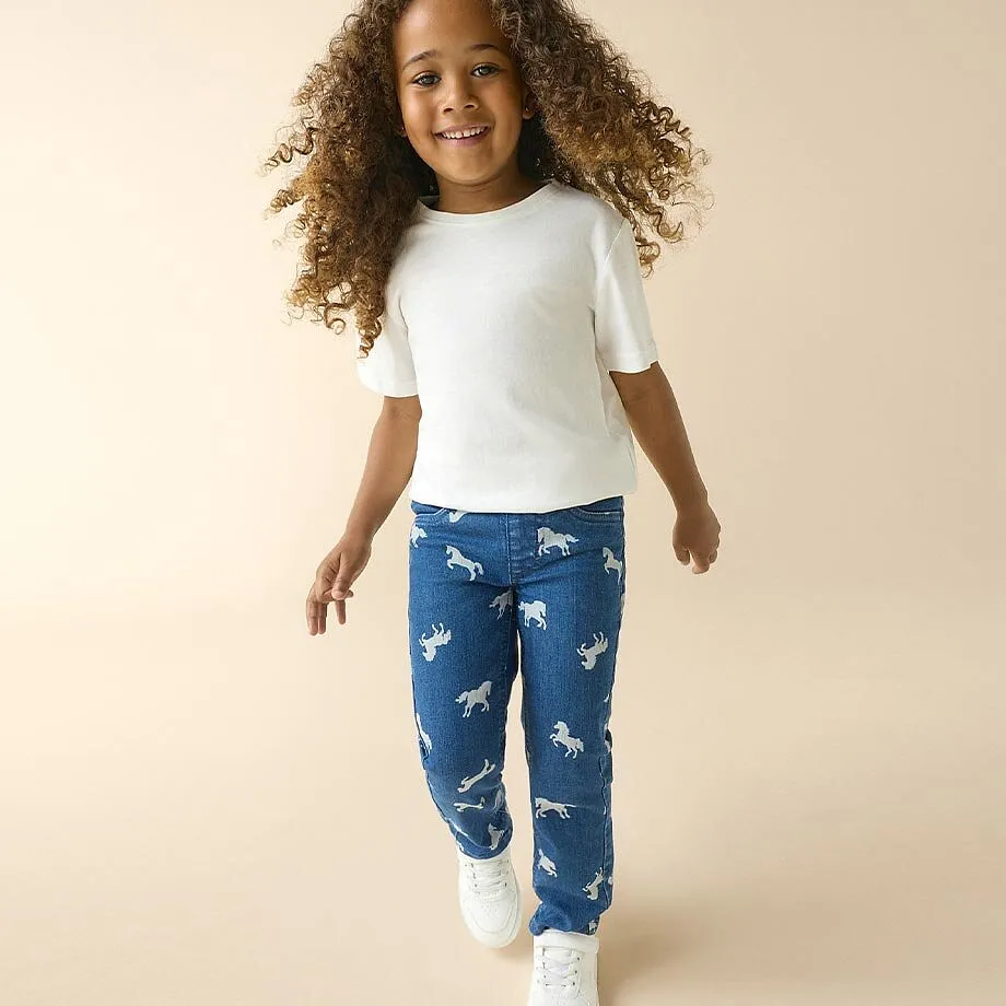 Flickan b&auml;r en vit T-shirt, bl&aring; jeans med h&auml;stm&ouml;nster och vita sneakers.