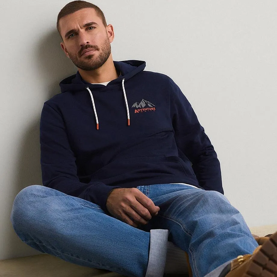Han har p&aring; sig en m&ouml;rkbl&aring; huvtr&ouml;ja med bergmotiv och ett par lediga, bl&aring;a jeans till det.