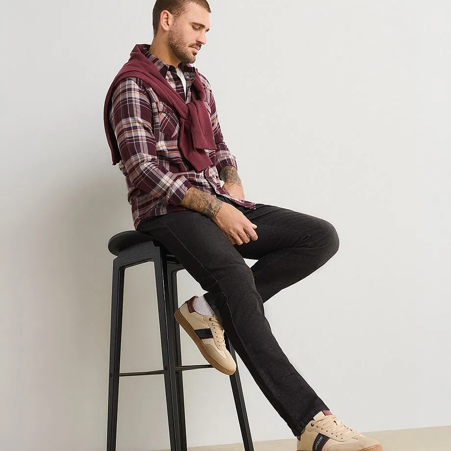 Mannen har p&aring; sig en rutig skjorta, m&ouml;rka jeans, beigea gymnastikskor och en vinr&ouml;d tr&ouml;ja.