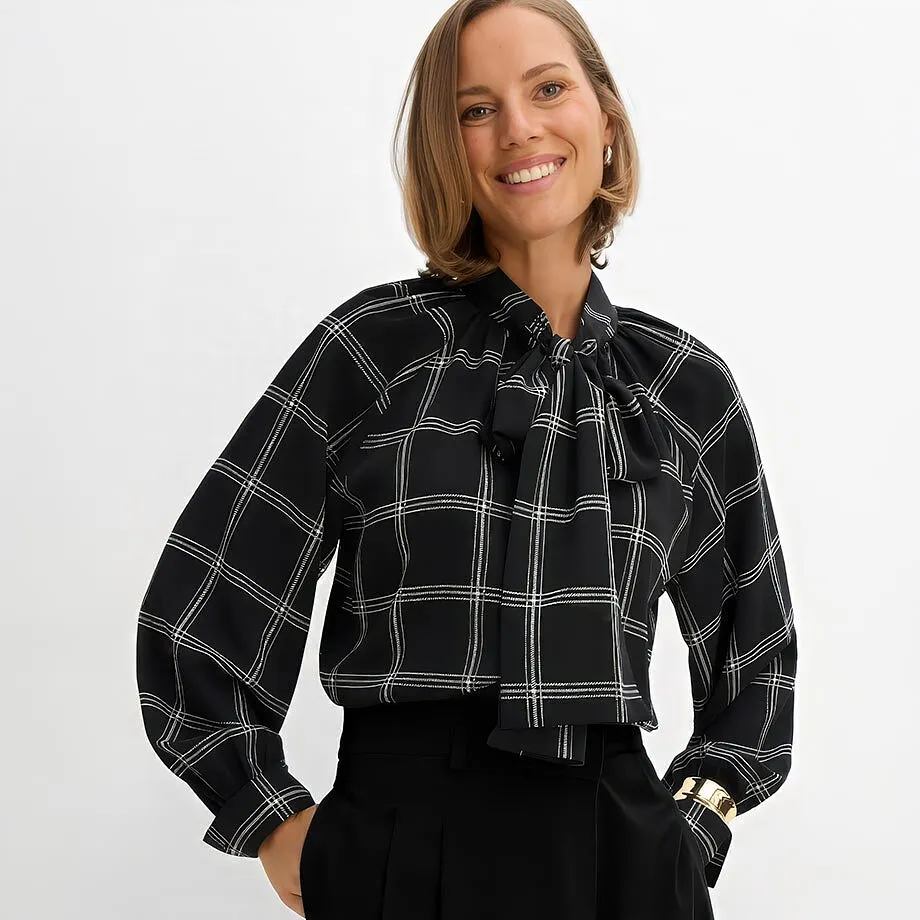 Elegant, svart blus med rutm&ouml;nster och knytband, svarta byxor, guldpl&auml;terat armband.
