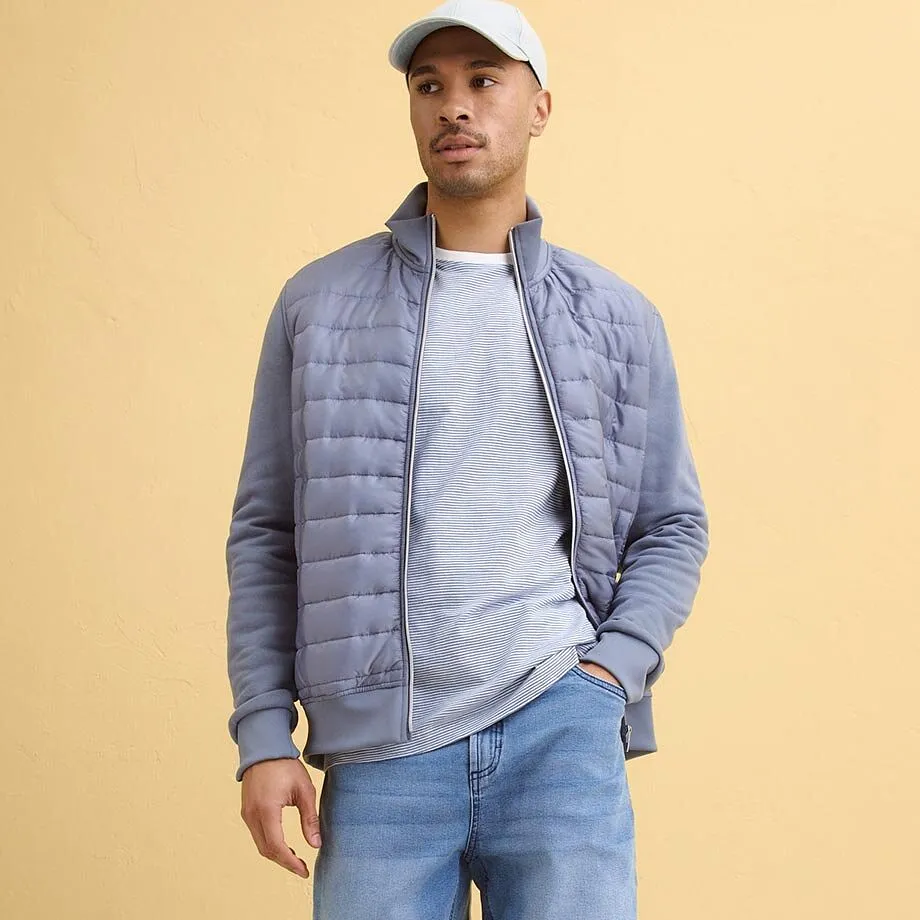 Gr&aring;bl&aring; &ouml;verg&aring;ngsjacka &ouml;ver ljusgr&aring; tr&ouml;ja, bl&aring; jeans.

